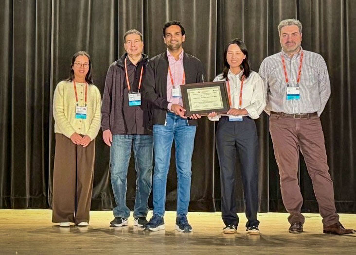 Five people stand on a stage with a dark curtain backdrop, the fourth holding a framed certificate.