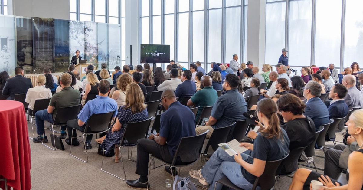 A speaker addresses a seated audience at the F3I Supercomputing Center launch inside a bright, modern hall.