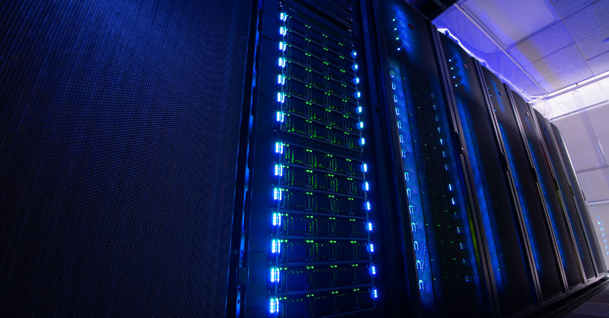 A row of Expanse server racks in SDSC's data center illuminated with glowing blue and green LED lights.