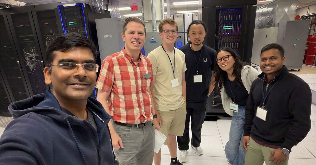 Six diverse adults pose for a selfie inside the SDSC server room with rows of computer racks in the background.