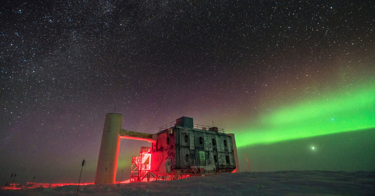 A research station sits in a snowy landscape under a starry night sky with a green aurora and glowing red lights.
