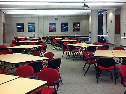 Interior view of the SDSC Auditorium in an angled table configuration, featuring pairs of rectangular tables with black and red chairs arranged for small group discussions.