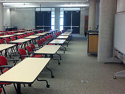 Interior view of the SDSC Auditorium in a classroom configuration, featuring rectangular tables each with three black and red chairs facing the front of the room.