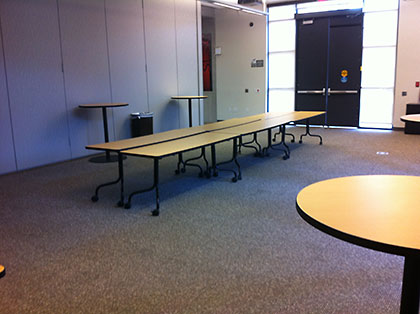 Folding tables and round cafe tables in a carpeted multi-purpose room with grey walls and double doors.