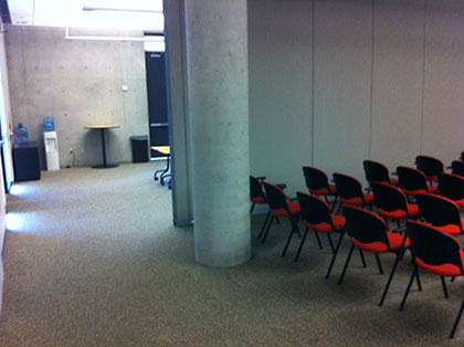 Back of the auditorium with rows of red and black chairs, a large concrete pillar, and a water cooler in a connecting open space.